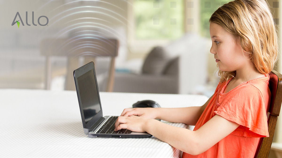 A young girl sits at a table using a laptop computer in a bright, modern home setting. The child is focused on the screen, with her hands on the keyboard, suggesting an activity such as learning or playing. The background is softly blurred, highlighting the subject, and the word 'Allo' is clearly visible in the upper left corner. The overall mood is calm and studious, with natural daylight illuminating the scene.