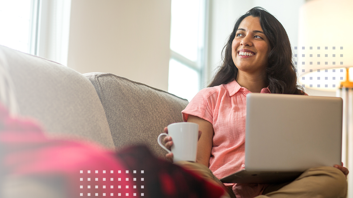 A woman sits comfortably on a sofa with a laptop on her lap and a coffee mug in hand