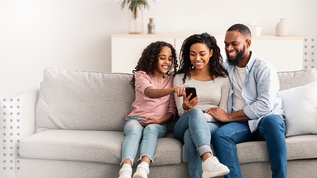A family of three sits closely on a light-colored sofa in a modern living room. The child is holding a remote control, possibly interacting with a television or device, while the adults sit on either side. The setting features soft, neutral tones and minimalistic decor, creating a cozy and inviting atmosphere.