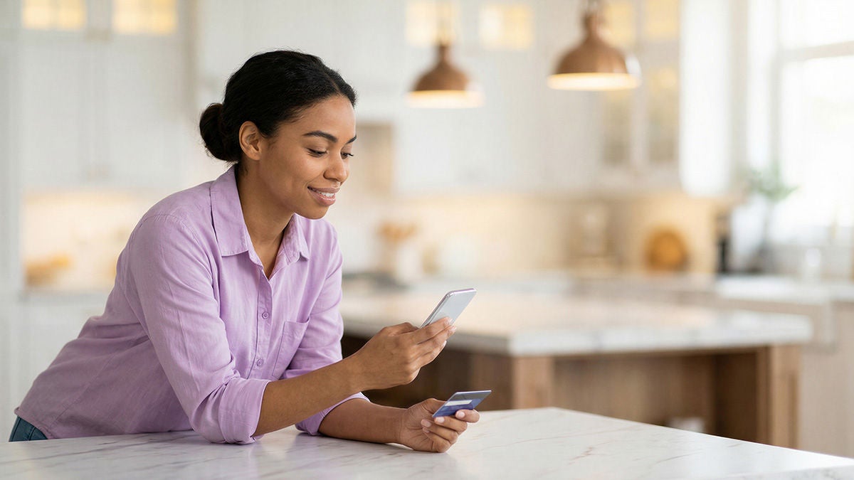 A young woman stands at a bright kitchen counter using her smartphone while holding a credit card. She appears focused and relaxed, suggesting online shopping or mobile banking. Warm pendant lights and a neutral, modern kitchen interior create a calm, inviting atmosphere. The scene emphasizes everyday digital payment and home lifestyle.