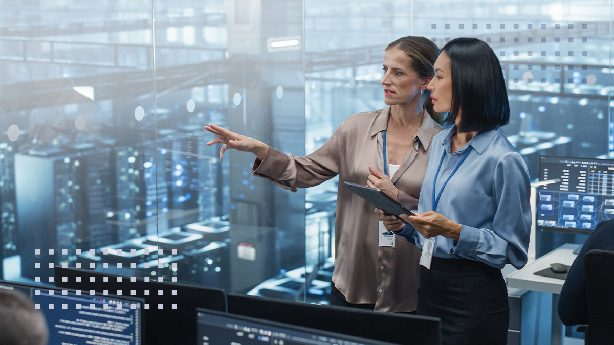 Two professional women stand in a modern control room, reviewing information on a tablet and multiple computer monitors
