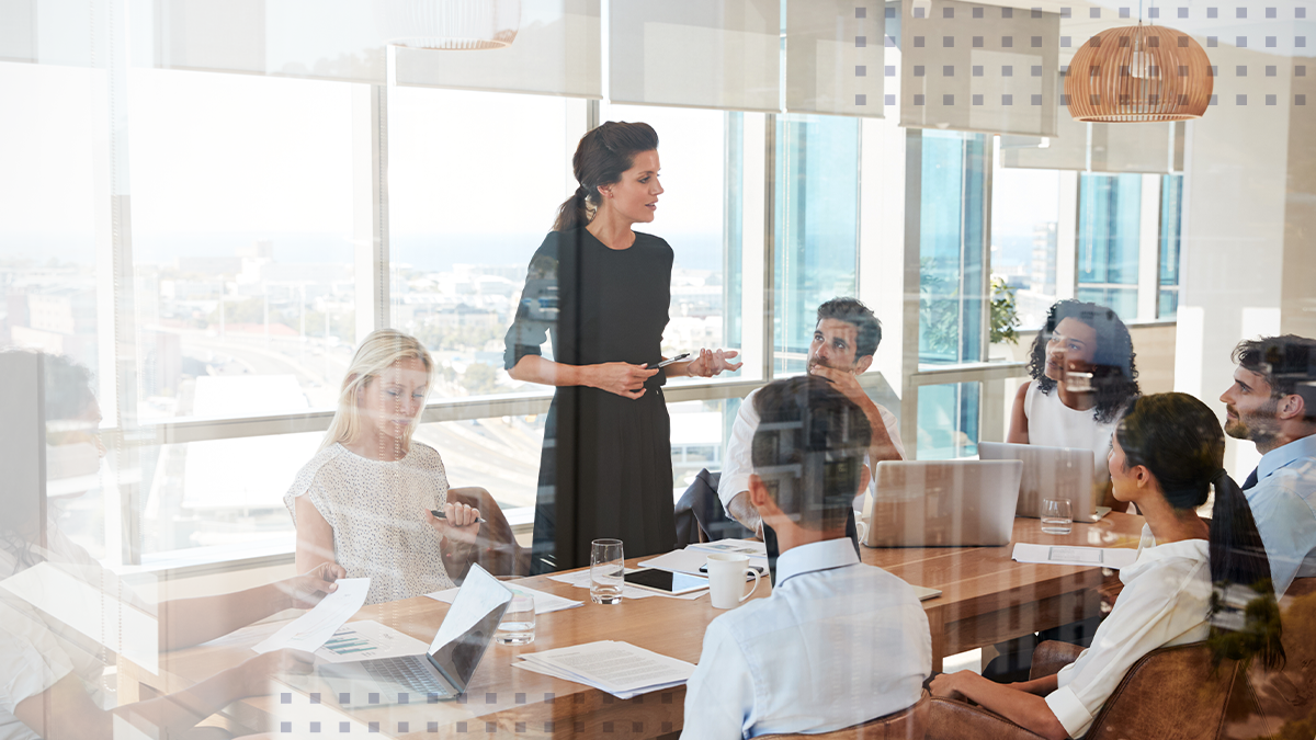 A group of employees sitting around a table in an office with one woman standing up and presenting