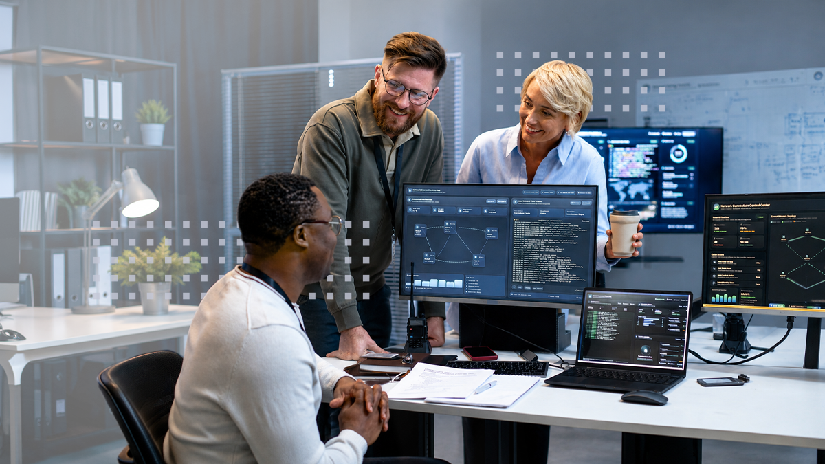 A smiling group of coworkers surrounding a desk with computer monitors displaying broadband operations AI data graphs and metrics