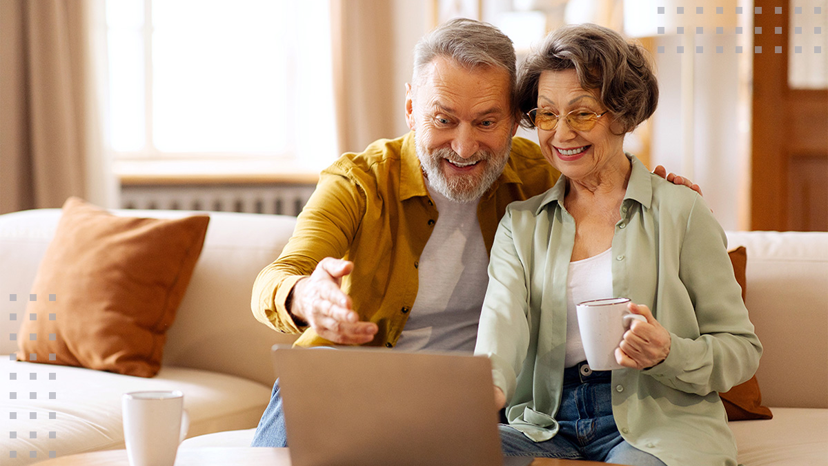 An older couple sits together on a couch, looking at a laptop screen while one person gestures toward it