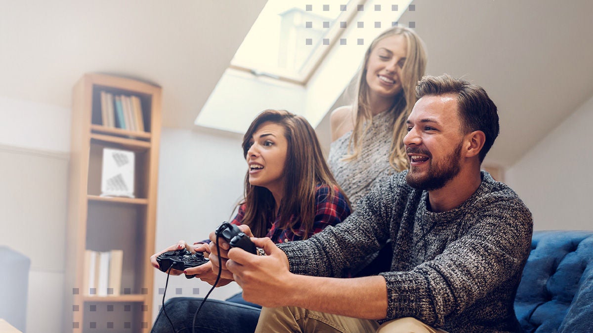 A group of three people sit closely together on a couch, focused on playing a video game with controllers in hand. They are in a cozy, modern living room with a bookshelf and slanted ceiling in the background. The image includes on-screen text elements such as a blue "PRESS RELEASE" banner and a visible WiFi 7 certification badge. The overall scene conveys home entertainment, connectivity, and shared leisure time.