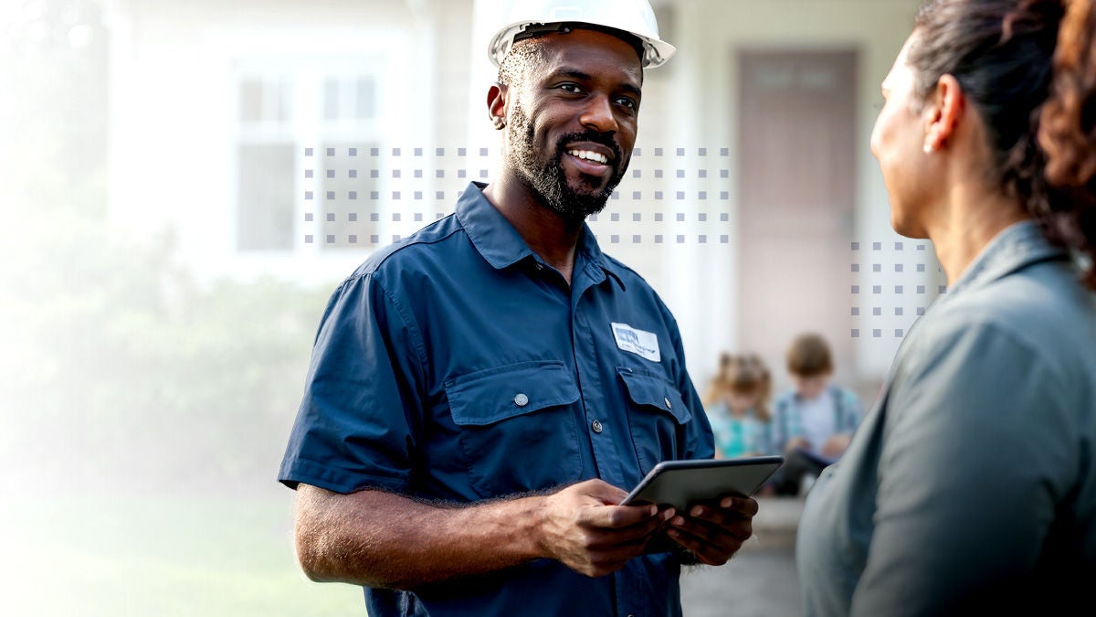 A male utility worker in a blue uniform and white hard hat stands outside a residential home, holding a digital tablet. He is engaged in conversation with a woman, possibly the homeowner, while two children are visible in the background near the entrance. The setting is a suburban neighborhood, and the mood is professional and approachable.