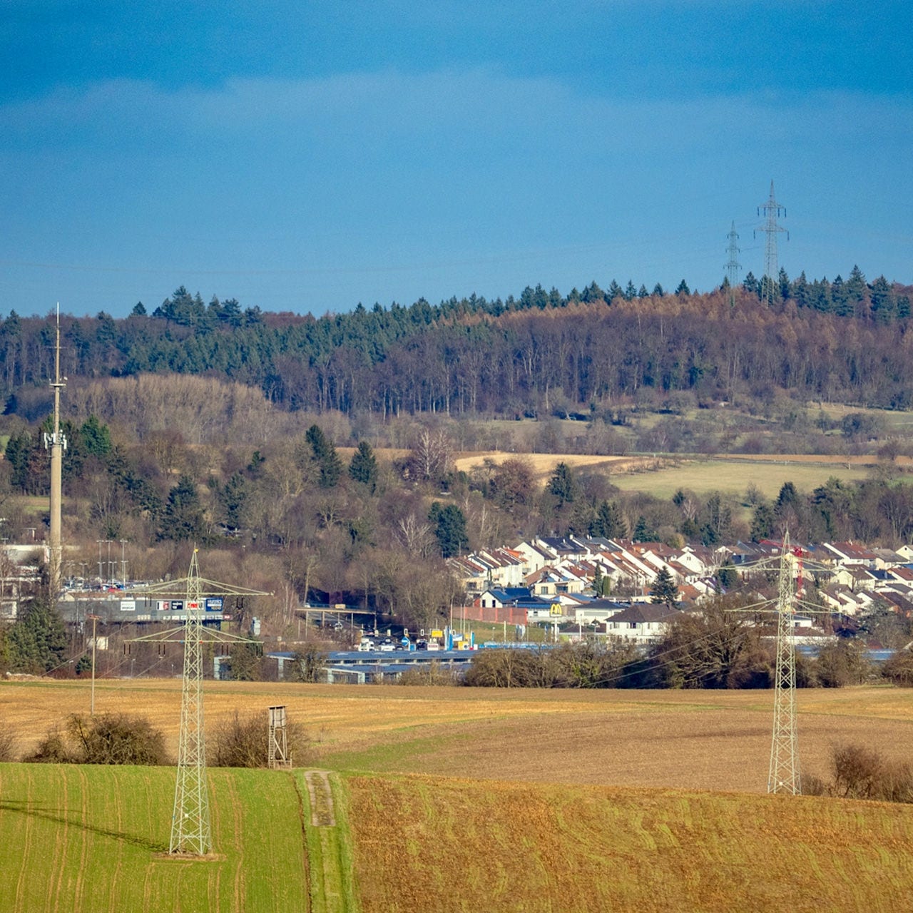 A suburban neighborhood with rows of houses sits at the edge of open agricultural fields. In the midground, utility poles and transmission towers stretch across the landscape, connecting the residential area to surrounding infrastructure. Behind the houses, a dense forested hillside rises, adding natural contrast to the built environment. The sky above is clear and blue, giving the scene a calm, early spring atmosphere.