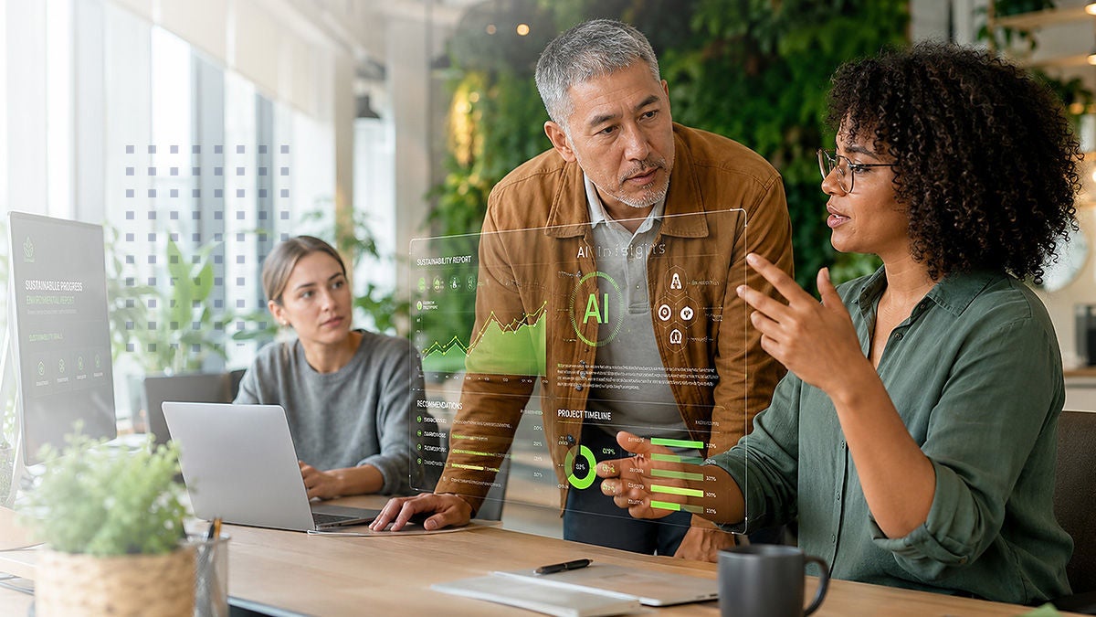 Three colleagues collaborate around a modern desk while viewing a transparent augmented reality data dashboard. A woman in the foreground explains information to a standing man, as another teammate listens from behind a laptop. The interface shows charts, graphs, and the letters AI in green, suggesting artificial intelligence analytics. Large windows and indoor plants create a bright, contemporary office environment.