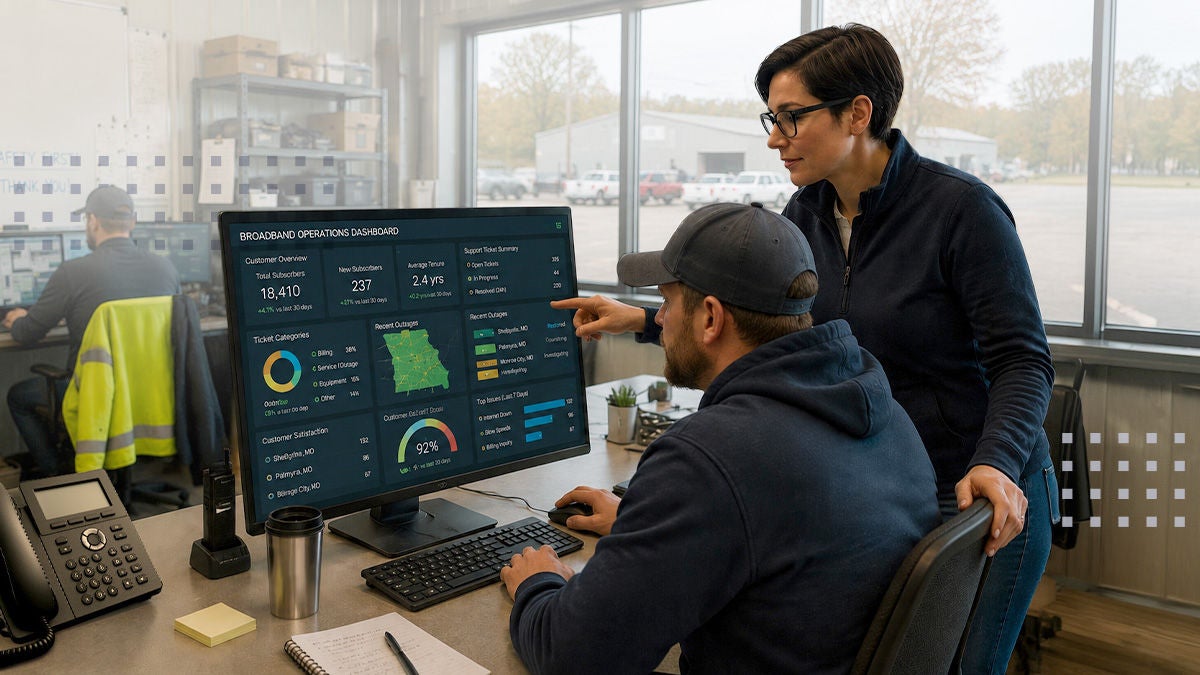 Two coworkers analyze a large data dashboard on a desktop monitor in a modern industrial office. The seated man uses the keyboard and mouse while the standing woman points at the screen, discussing performance metrics and charts. The workspace includes phones, paperwork, and shelves with equipment, with another worker visible through the glass partition. The scene suggests data-driven decision making and collaboration in a professional environment.