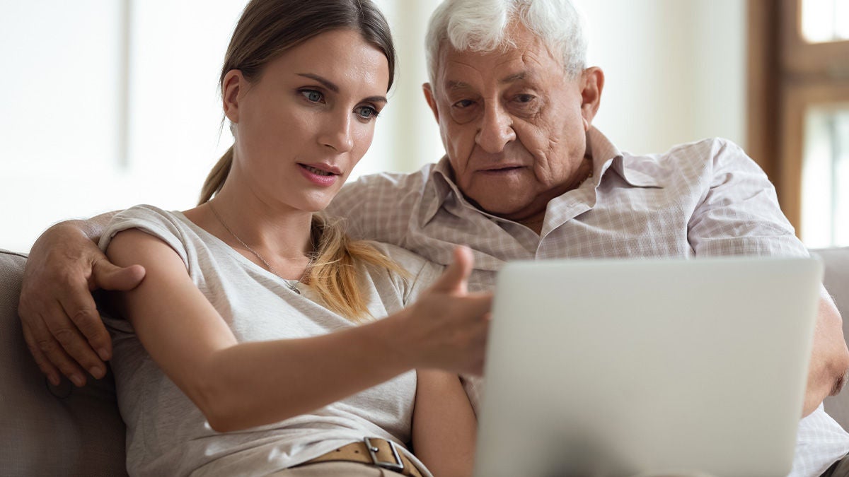 A young woman sits closely with an older man while they look at a laptop screen together. She gestures toward the screen, explaining or demonstrating something as he listens attentively. They sit on a comfortable couch in a bright indoor setting, suggesting a home environment. The scene conveys intergenerational support, learning, and family connection.