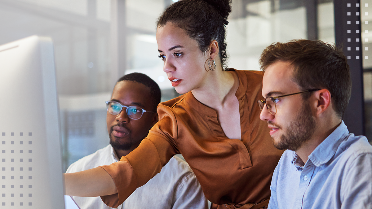 A woman with a serious facial expression pointing at a computer monitor beside two male coworkers on a marketing team