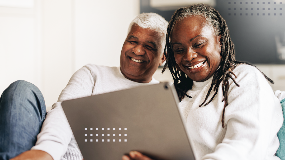 An older couple sits closely together on a couch, smiling as they look at a large tablet screen