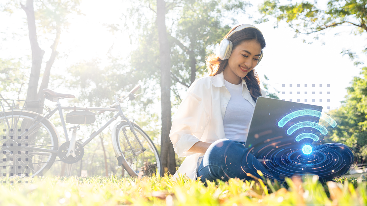 A woman with headphones on sitting in a grassy park area with a laptop with an abstract Wi-Fi symbol overlaid