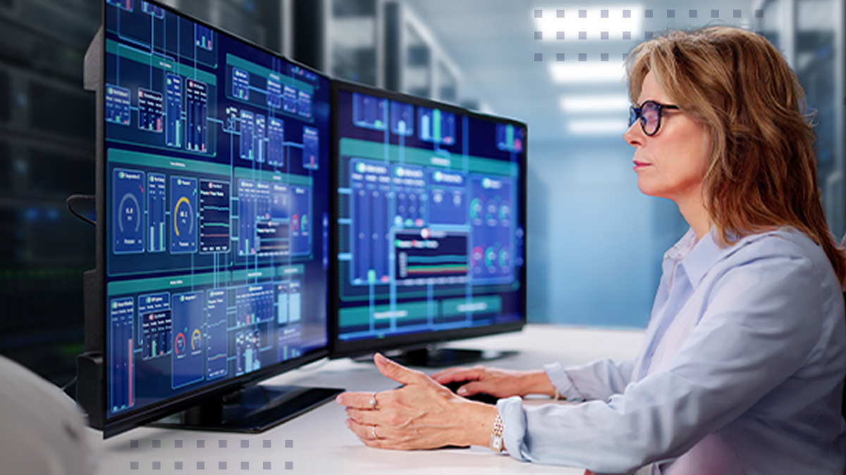 A professional sits at a workstation monitoring two large computer screens filled with complex network operations dashboards