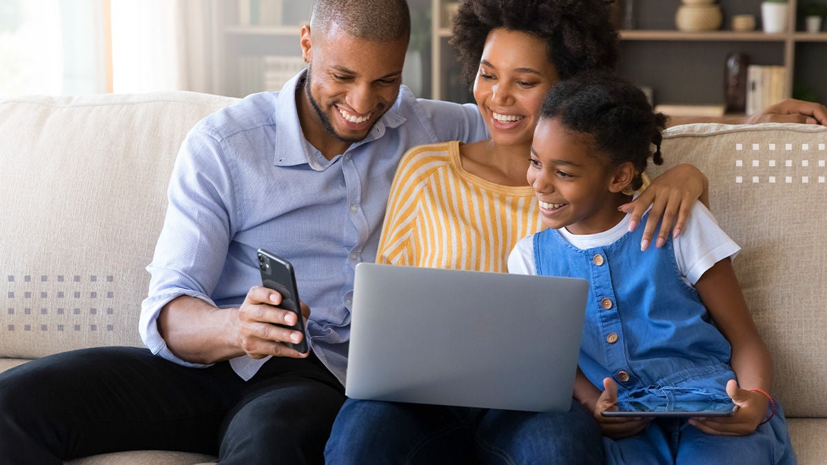 A smiling family sits closely together on a couch, enjoying time with digital devices. The father holds a smartphone while the mother uses a laptop on her lap. A young child sits beside them, looking at a tablet and engaging with the parents. The bright living room setting and relaxed posture convey warmth, connection, and modern home life.