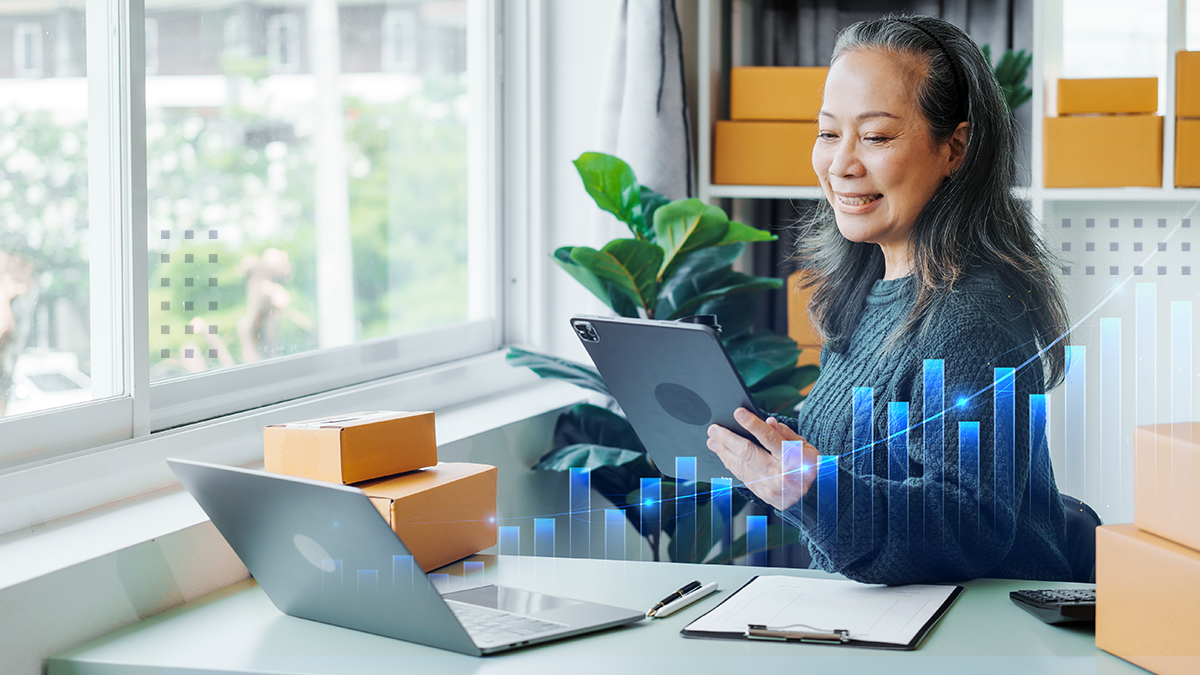 A smiling woman in an office using a tablet and laptop at a desk with a positive trending abstract graph overlaid