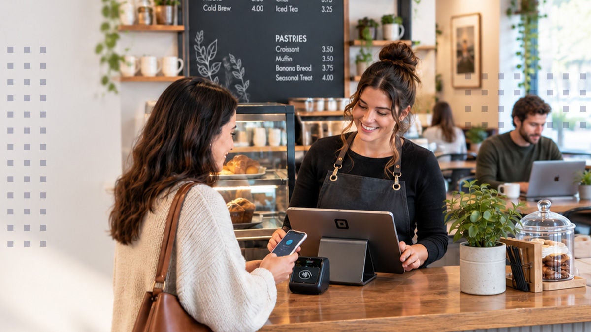 A smiling barista stands behind a wooden counter, assisting a customer who is paying with a smartphone at a small card reader. The scene takes place in a modern, cozy café with a pastry display case and a large chalkboard menu in the background. Warm lighting, indoor plants, and seated patrons create a welcoming atmosphere. The focus is on digital payment, friendly service, and a relaxed coffee shop environment.