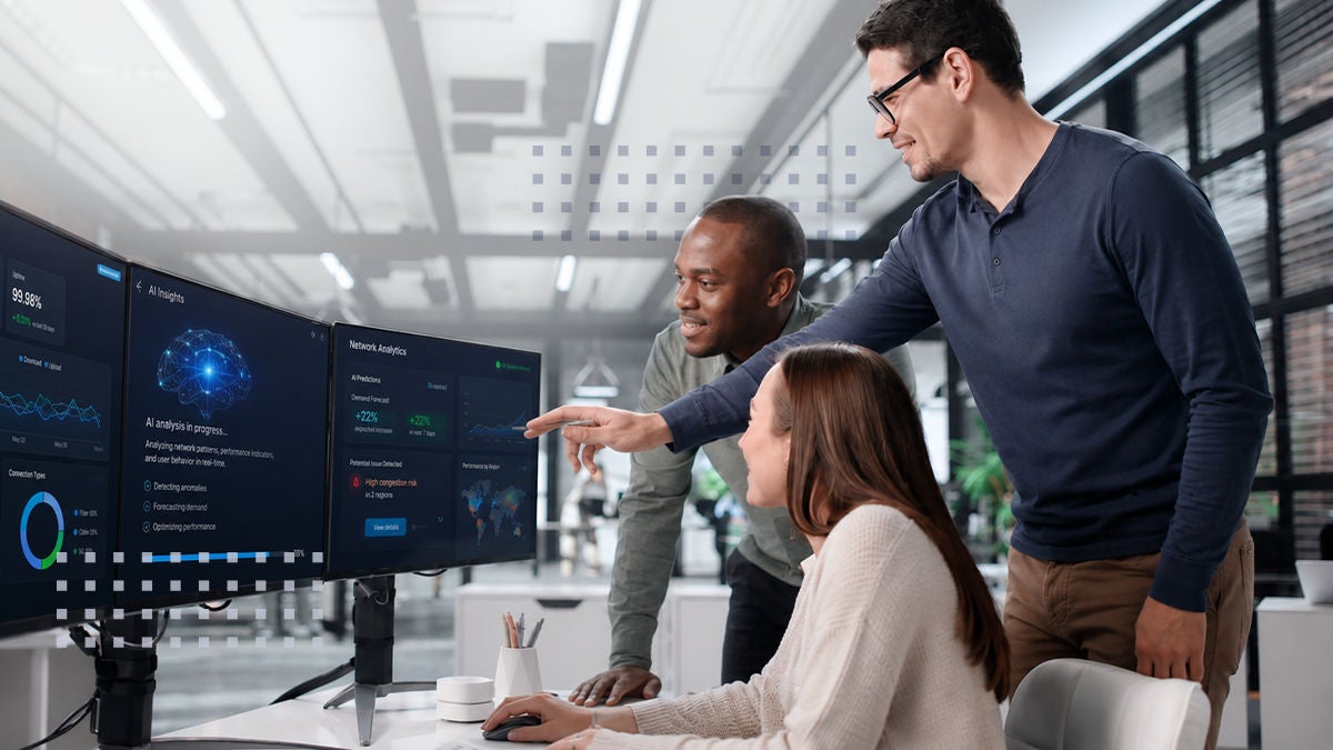 Three coworkers collaborate in a modern open-plan office, focused on large dual monitors displaying data analytics dashboards. 