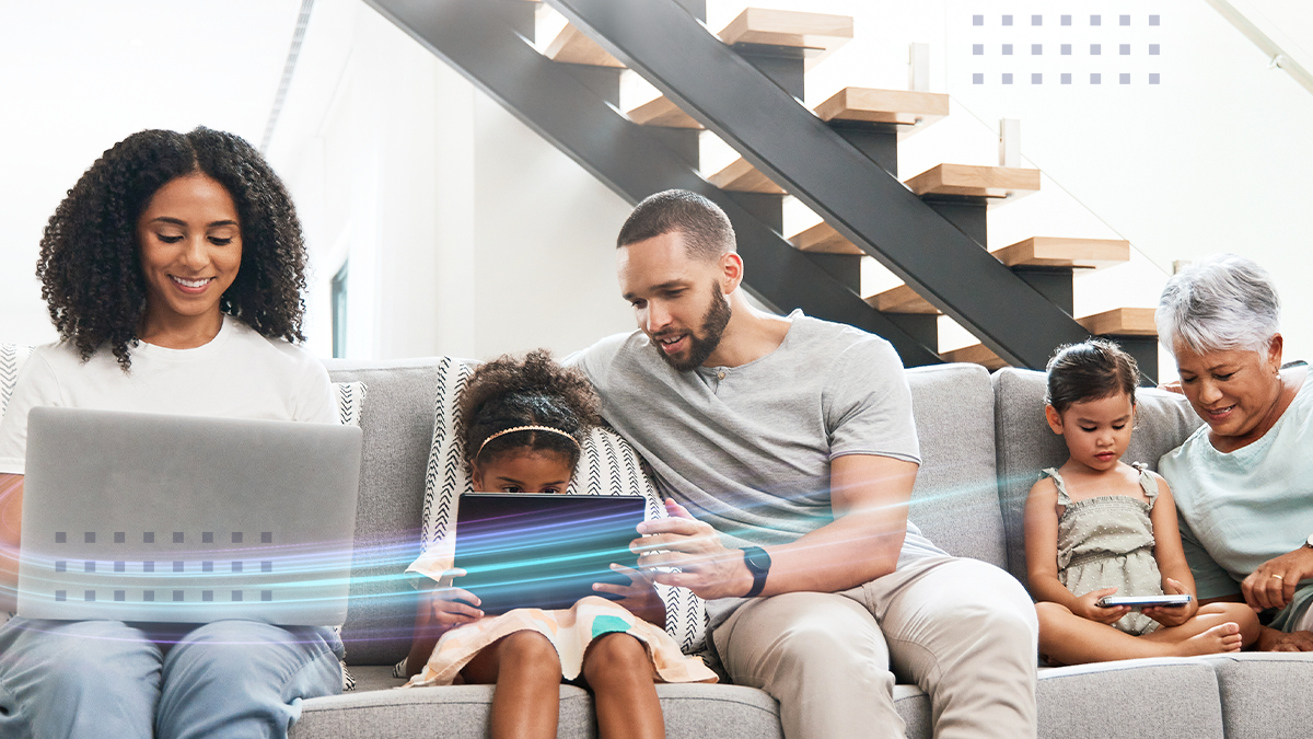 A diverse family sits together on a sofa in a bright, modern living room, each person engaged with a digital device such as a laptop or tablet