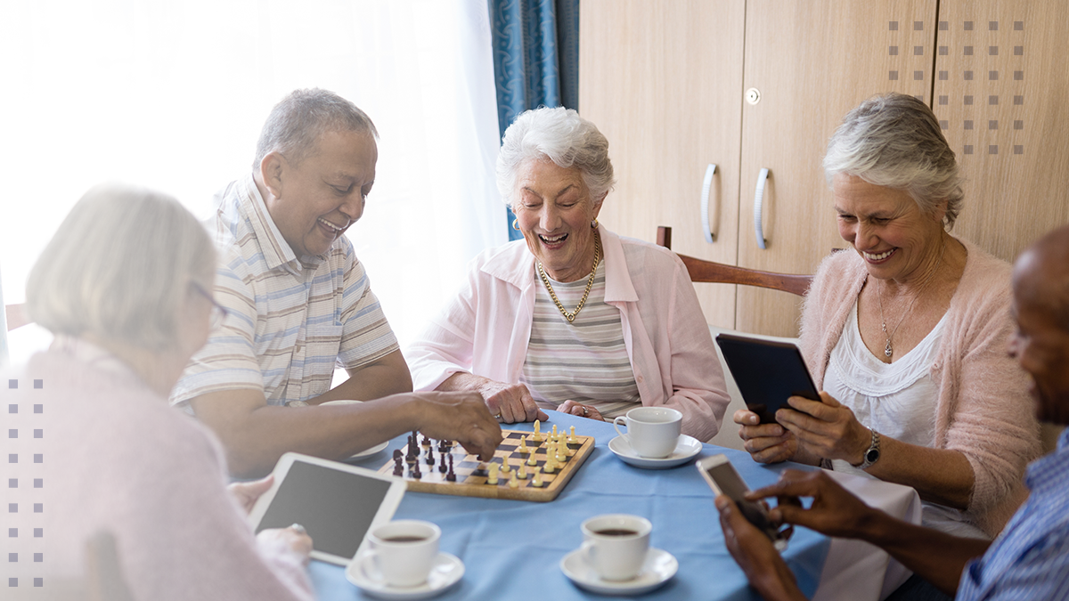 A group of older adults sit together at a table, enjoying a game of chess while others use digital tablets