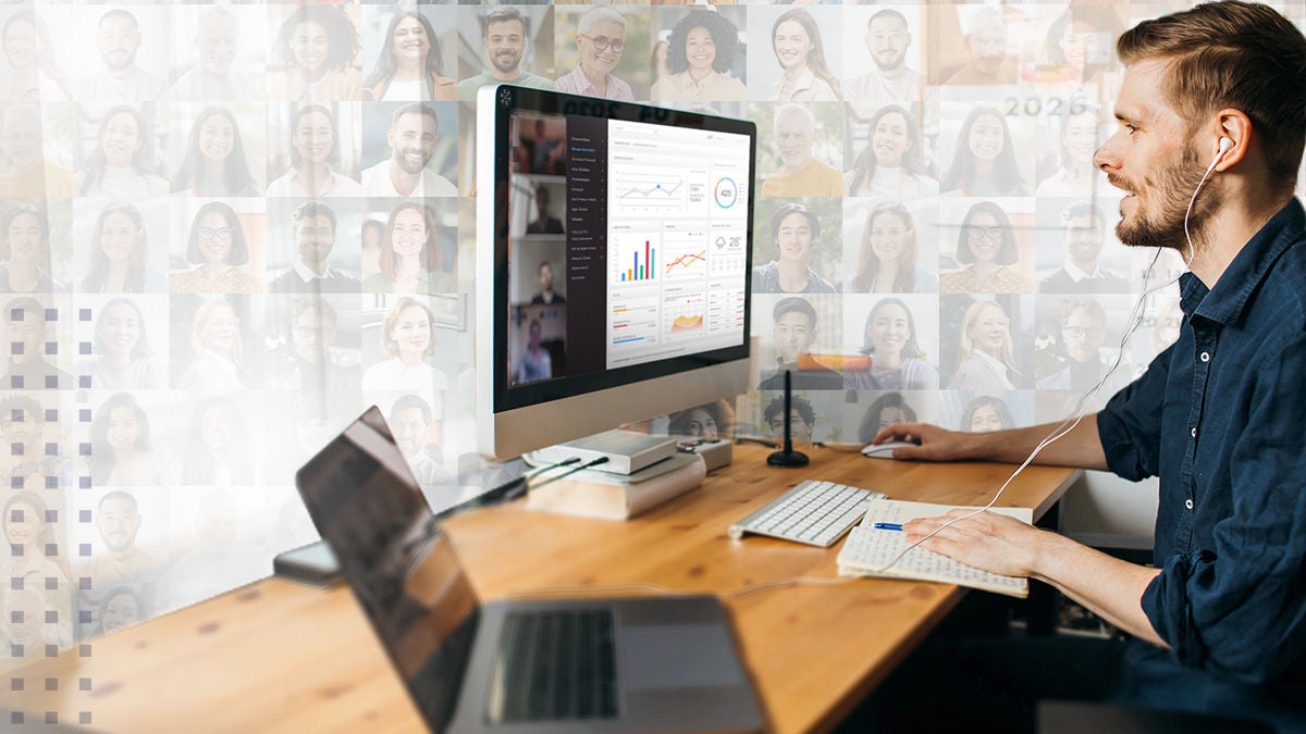A young adult male sits at a wooden desk, engaged in remote work on a desktop computer. The screen displays a video conference interface with charts, graphs, and multiple participant windows. The workspace includes a laptop, notebook, and headphones, suggesting a professional and focused environment. The background features a collage of diverse faces, emphasizing virtual collaboration.