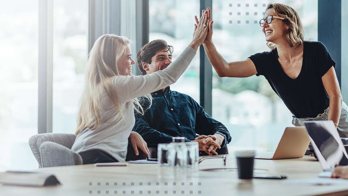Three colleagues sit together in a modern office, enthusiastically high-fiving across a conference table