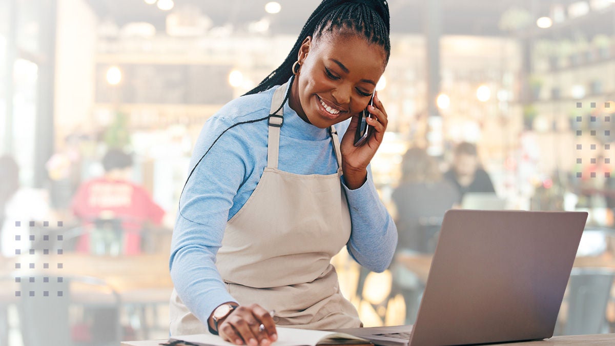 A person wearing a beige apron and blue shirt is seen working in a cafe environment. They are using a laptop while talking on the phone, suggesting multitasking or managing business operations. The background shows a bright, modern cafe with blurred patrons and tables. The overall mood is busy and professional, with natural lighting and a warm palette.