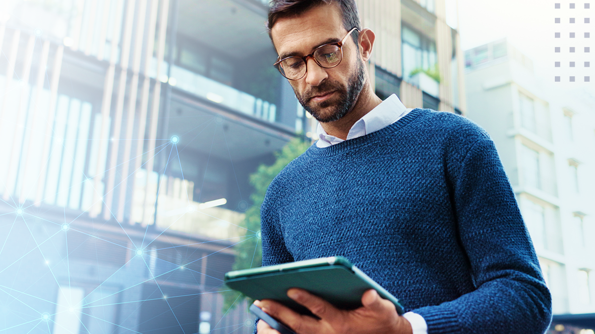 A man wearing glasses and a blue sweater looks down at a tablet device in his hands