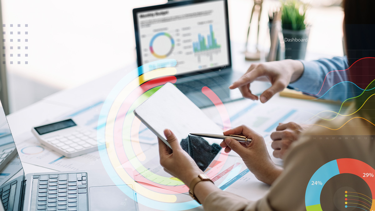 Two people collaborate at a modern office desk, reviewing data on a tablet and laptop